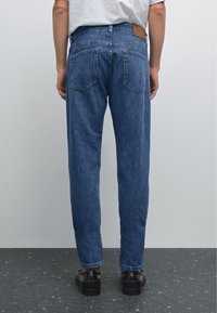 Jean en denim bleu clair avec une coupe droite, cinq poches, un patch en cuir à l'arrière et des coutures contrastées. Porté avec des chaussures noires.