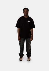 Černé oversized tričko s bílým textem "CRYSTAL", v kombinaci s tmavými denimovými džíny a světlými teniskami. Model stojí čelem.