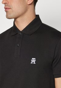 Polo nero realizzato in misto cotone. Presenta un colletto a costine, una patta con due bottoni e un logo ricamato bianco sul petto.