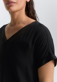 Blouse noire avec un décolleté en V et des manches courtes retroussées ; fabriquée en tissu lisse avec un léger éclat ; présente un détail de décolleté transparent.