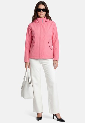 Vrouw met een roze capuchonjas, witte broek, zwarte hakken, zonnebril en een witte handtas, tegen een effen achtergrond.