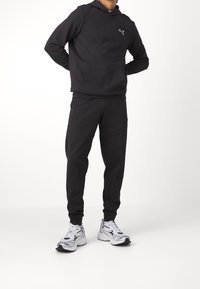 Conjunto de sudadera con capucha y pantalones jogger negros hechos de una mezcla de algodón. La sudadera incluye un bolsillo canguro y puños acanalados. Combinado con zapatillas blancas y grises.