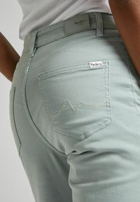 Light green denim jeans with a fitted design, featuring back pockets and subtle wave stitch detail. Includes a small logo tag.