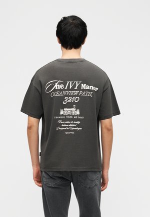 Άνδρας που φοράει σκούρο γκρι μπλουζάκι με λευκό κείμενο και εικόνα στο πίσω μέρος που γράφει "The Ivy Manor, Oceanview Path, 3210".