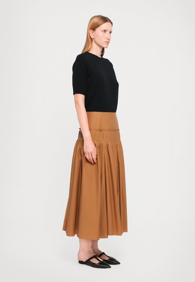 Schwarzes kurzärmliges Oberteil, kombiniert mit einem braunen plissierten Midirock. Das Model trägt schwarze Slippers, und das Outfit besticht durch ein klares, modernes Design.