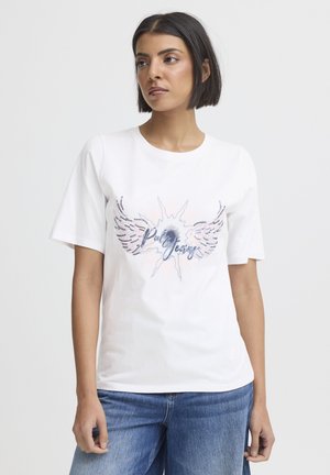 Kvinde iført en hvid T-shirt med vinge- og solstrålegrafik samt teksten "Polo Jeans", båret sammen med blå jeans, stående foran en ensfarvet baggrund.