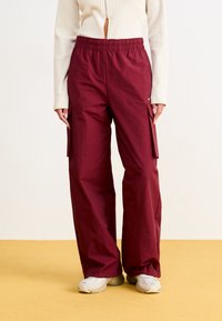 Burgunderfarbene Cargo-Hosen mit elastischem Bund, Seitentaschen, lockerer Passform und glatter Textur. Kombiniert mit hellen Schuhen.