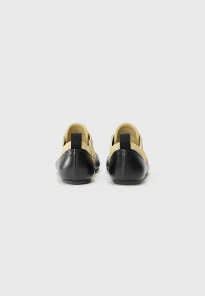 Zapatos de cuero negro y beige con puntera redondeada, con un tacón bajo y un lazo en la parte trasera para facilitar su puesta. Textura suave y diseño minimalista.
