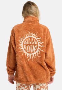 Veste en polaire orange duveteuse avec un col haut, pr�ésentant un grand motif de soleil blanc au dos. Coupe décontractée et oversize.