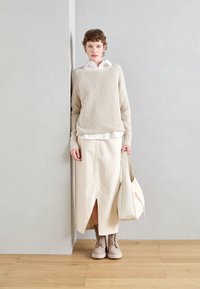 Beige neulepusero valkoisen paidan päällä, kermankeltainen kietaisuhame, jossa on halkio, beige nauhakenkä, ja valkoinen käsilaukku, vaaleaa seinää vasten.