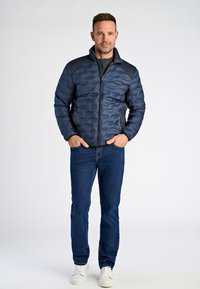 Marinblå pufferjacka med kviltad textur, solida axelpaneler, två sidofickor, kombinerad med blå jeans och vita sneakers.