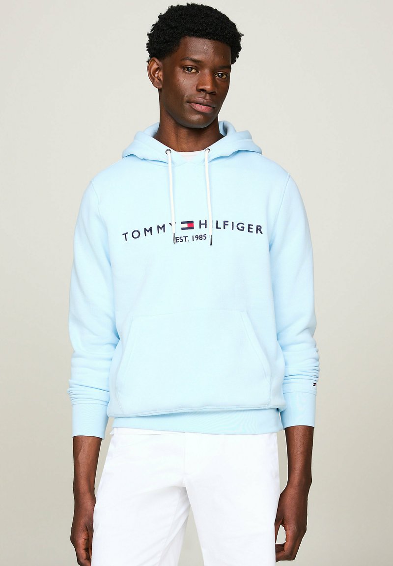Sudadera azul clara de algodón, con un bolsillo frontal tipo canguro, capucha con cordones y logotipo impreso en azul marino y rojo. Pantalones blancos.