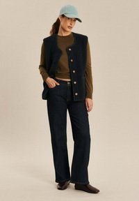 Marine fleece vest met vier knopen, bruine langemouwen shirt, donker wijde jeans en lichtblauw petje. Bruine schoenen maken de outfit compleet.