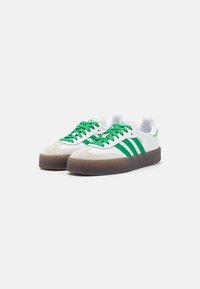 Baskets blanches avec accents verts et trois bandes, dotées d'un capuchon en cuir suédé, d'une semelle en caoutchouc texturée et de lacets verts.
