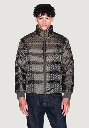 Ung mand iført en sort dunjakke med et gentaget "AX"-mønster og mørke jeans, stående foran en ensfarvet lys baggrund.