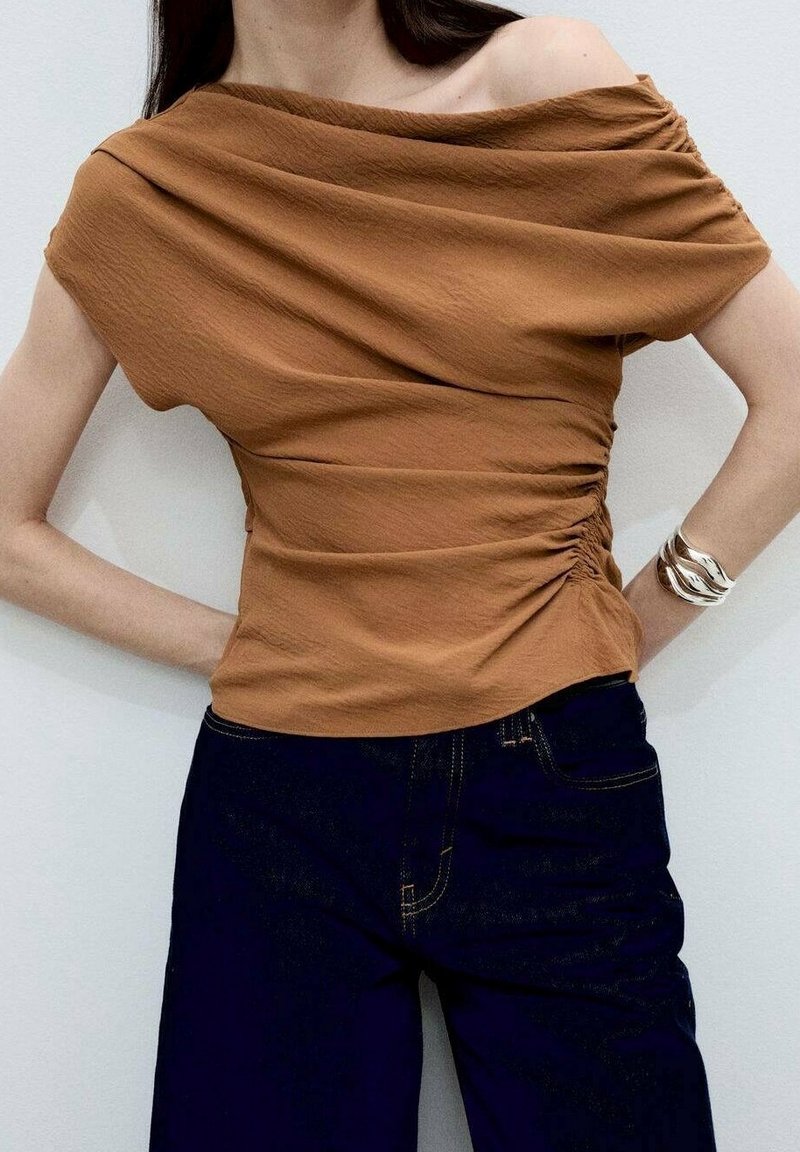 Brun, off-shoulder bluse med tekstureret stof og samlet sideskæring, parret med mørk denim jeans med gul syning.