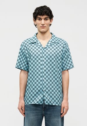 Noor mees, kes kannab helesinist lühikeste varrukatega nööbitavat särki musta ruudulise mustriga ja siniseid teksapükse, seisab tavalise tausta ees.