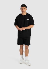 Svart bomullst-shirt med avslappnad passform och korta ärmar, matchad med svarta shorts. Vita skor och strumpor med logodetaljer kompletterar outfiten.