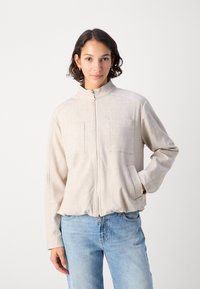 Giacca zip-up beige chiaro con colletto alto, due tasche sul petto e orlo elasticizzato. Presenta un tessuto testurizzato in una vestibilità rilassata.