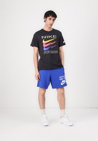 Tricou sport Nike negru cu un logo colorat, asortat cu pantaloni scurți albaștri ce au text alb. Sneakers albi și șosete de culoare deschisă completează ținuta.