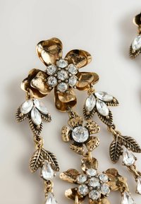 Ohrringe in goldfarbener Ausführung mit floralen Motiven, ausgestattet mit strukturierten Metallblumen und Blattakzenten, verziert mit klaren Strasssteinen in unterschiedlichen Größen.