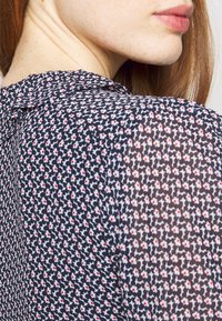 Gros plan sur le cou et l'épaule d'une femme portant un chemisier noir avec de petites fleurs blanches aux centres rouges, montrant une peau lisse et des cheveux roux.