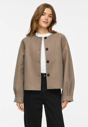 Femme portant une veste boutonnée taupe sur un haut blanc avec un pantalon noir sur un fond clair uni.