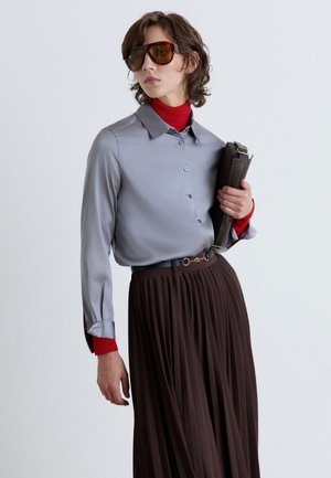 Person wearing oversized brown sunglasses, gray button-up shirt over a red turtleneck, dark pleated skirt, holding a black clutch bag.