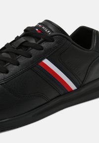 Basket en cuir noir avec des lacets noirs, comportant un détail avec des bandes rouge, blanche et bleue ainsi que le nom de la marque embossé sur le panneau latéral.