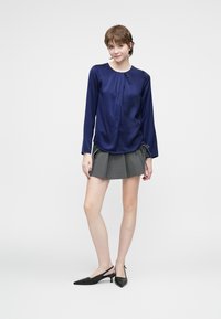 Giovane donna in piedi, che indossa una blusa blu navy a maniche lunghe, una mini gonna plissettata grigia e delle décolleté nere con tacco kitten su uno sfondo bianco.
