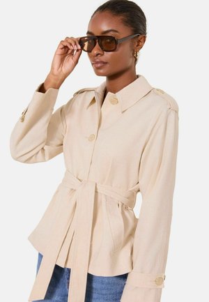 Frau, die eine große braune Sonnenbrille justiert, trägt eine beige Jacke mit Gürtel und Knöpfen sowie blaue Jeans, vor einem einfarbigen Hintergrund.