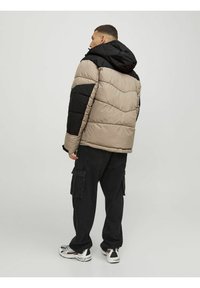 Giubbotto imbottito beige e nero, con cappuccio e design trapuntato. Indossato con pantaloni cargo neri e sneakers bianche e nere.