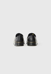 Black leather shoes with a sleek design, rounded shape, and textured soles. Features yellow stitching along the base. Rear view.