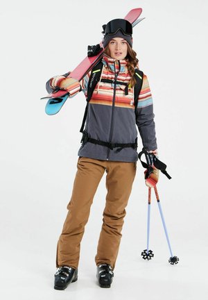 Skibekleidung mit einer mehrfarbigen gestreiften Jacke, braunen Hosen, schwarzen Skischuhen und pinkfarbenen Skiern mit blauen Stöcken und Griffen.