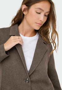Bruine blazer van gestructureerde stof met een notch-kraag, enkele knopsluiting en een voorzak. Draagt over een wit t-shirt.