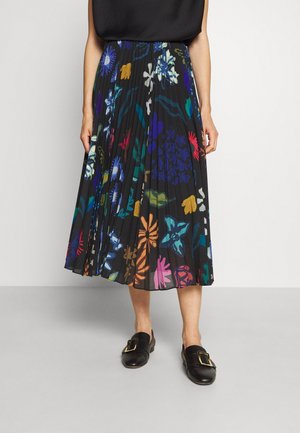 Femme portant une jupe midi noire plissée avec des motifs floraux colorés, un haut noir et des mocassins noirs à boucle, debout sur un sol gris.