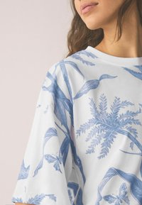 Valge t-särk suurendatud lõikega, millel on sinine botaaniline muster, mis sisaldab linde, lehti ja liblikaid, valmistatud pehmest kangast.