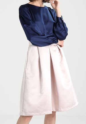 Mujer con blusa de satén azul marino de manga larga y falda midi de talle alto en línea A de color rosa pálido, sobre un fondo liso.