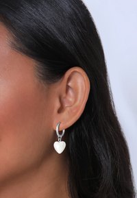 Boucles d'oreilles en argent en forme de hoop avec un charme en forme de cœur blanc, présentant une surface lisse et une finition polie, portées sur l'oreille d'un mannequin.