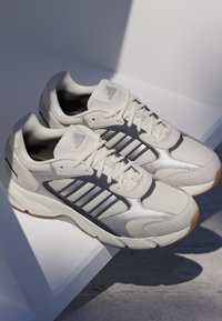 Białe sportowe sneakersy z szarymi akcentami, wykonane z siateczki i materiałów syntetycznych, o teksturowanej powierzchni oraz gumowej podeszwie z wzorem zapewniającym przyczepność.