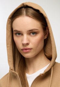 Felpa con cappuccio in tessuto beige, con chiusura a zip, dalla texture morbida e dal taglio classico, abbinata a una maglietta bianca sotto.