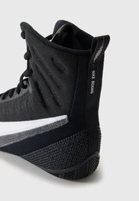 Boxerská obuv v černé síťovině s texturovanou podrážkou. Má bílý pruh přes střední část a branding "NIKE BOXING" na patě.
