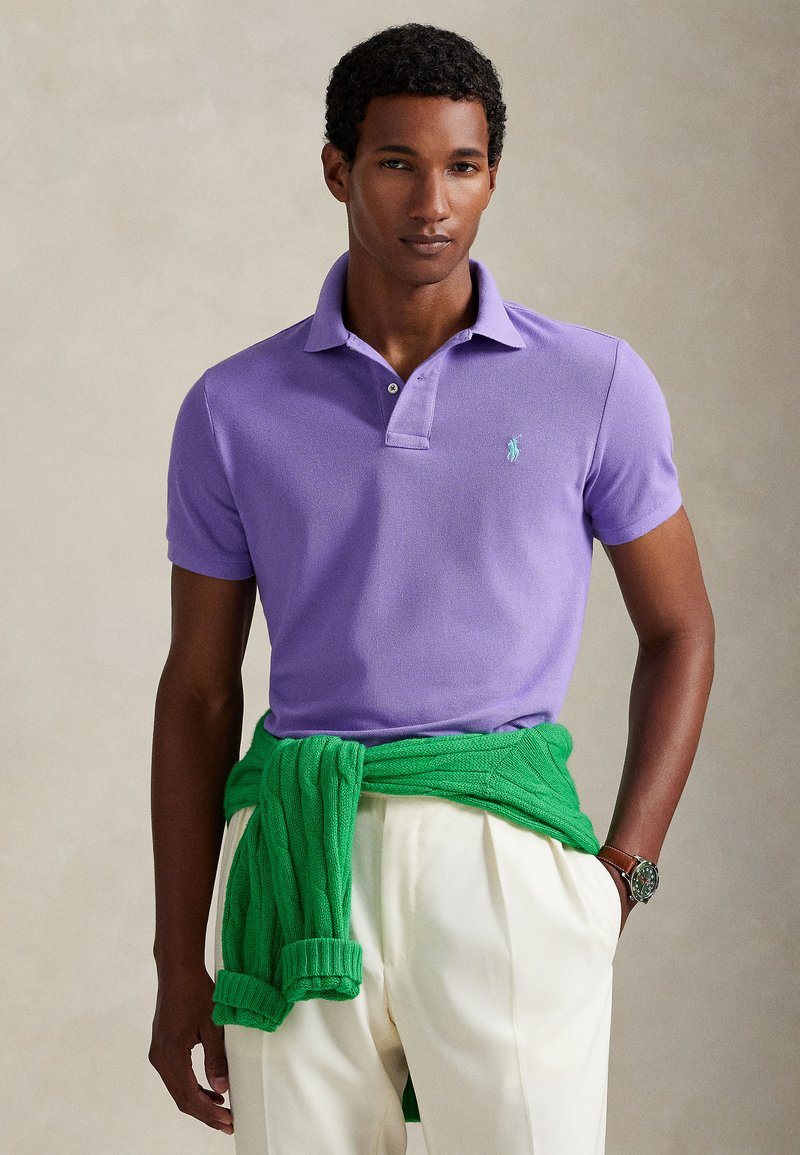 Mann trägt hellviolettes Poloshirt, weiße plissierte Hose, grünen Pullover um die Taille gebunden und eine braune Armbanduhr, posiert vor neutralem Hintergrund.