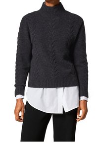 Personne portant un pull à col roulé gris foncé en maille torsadée par-dessus une chemise blanche avec un pantalon noir, debout les mains détendues.