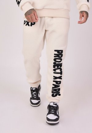 Joggers de felpa crema con "PROJECT X PARIS" en texto negro a lo largo del costado; combinados con zapatillas en blanco y negro. Tejido texturizado, puños elásticos.