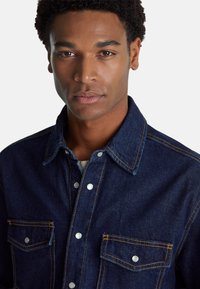 Camicia di jeans blu scuro con colletto, dotata di due tasche frontali, bottoni a pressione argentati e cuciture gialle a contrasto.