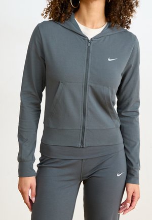 Femme portant un sweat à capuche zippé gris Nike et des leggings assortis, tous deux avec des logos Nike blancs, debout les mains détendues le long du corps.