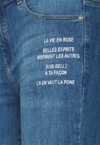 Vaqueros de mezclilla azul oscuro con texto bordado en blanco en el bolsillo trasero con citas inspiradoras en francés. Diseño clásico de cinco bolsillos.