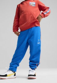 Kind trägt einen roten Puma Hot Wheels Hoodie, eine blaue Puma Hot Wheels Jogginghose und schwarz-weiße Sneaker mit gelben Akzenten, steht mit den Händen in den Hüften.