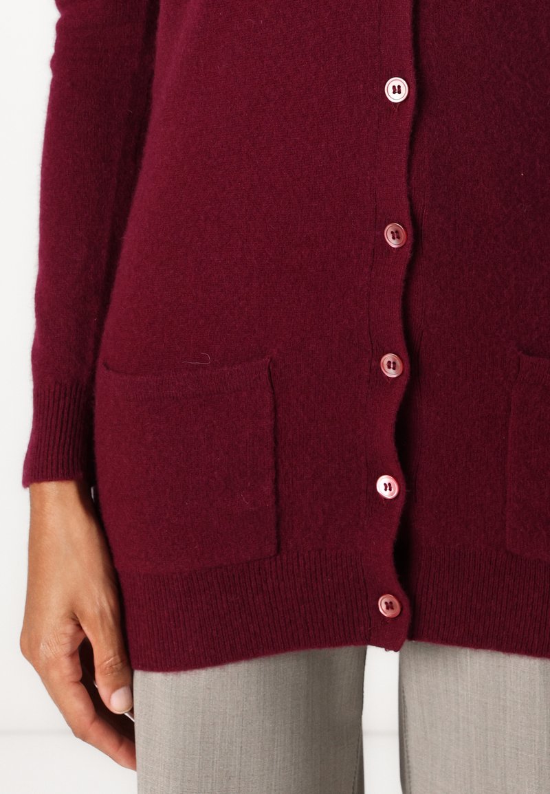 Burgunderfarbener Strickcardigan mit Knopfverschluss, zwei Taschen und gerippten Bündchen. Der Stoff wirkt weich und strukturiert.